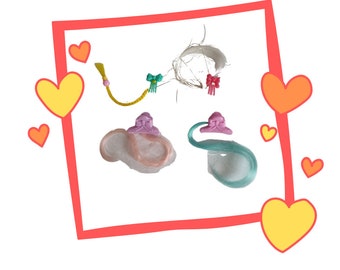 Könnte beinhalten: Eine Sammlung von Puppen-Haaraccessoires, darunter ein gelber Zopf, ein weißes Haarteil mit rosa Schleifen und zwei Haarteile mit rosa und blauem Haar. Die Accessoires sind vor einem weißen Hintergrund mit rotem Rahmen und Herzdekorationen.