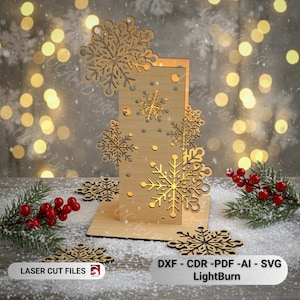 Può includere: Decorazione natalizia in legno tagliata al laser con disegni di fiocchi di neve. La decorazione ha una base rettangolare con un pannello verticale adornato con vari ritagli di fiocchi di neve. Il testo "LASER CUT FILES" e "DXF - CDR - PDF - AI - SVG LightBurn" è incluso.