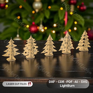 May include: Five laser-cut wooden Christmas tree decorations. Each tree is a light brown color with intricate cut-out designs, including snowflakes and stars. The trees stand on a dark surface with a blurred Christmas tree background.