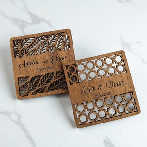 May include: Two square wooden coasters with intricate cut-out designs. One features a leaf pattern, the other a geometric design. Each coaster is engraved with a name and date: "Amelia and Ethan 18.08.2025" and "Lisa & Dean 25.08.2025".
