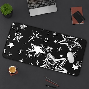 May include: Black desk mat with a white star pattern. The rectangular mat features a repeating design of various star shapes in a hand-drawn style. A computer mouse rests on the mat, alongside a laptop and other desk accessories.