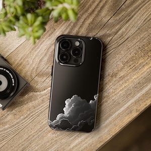 May include: Black smartphone case with a cloud design. The case features a glossy finish and a detailed illustration of white and gray clouds against a black background. The phone case is resting on a wooden surface.