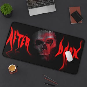 May include: Black rectangular mousepad with a red skull design and the words "AFTER DARK" in red flame-like lettering. A white mouse is on the right side of the mousepad. A laptop, a small plant, and a cup of coffee are also visible.