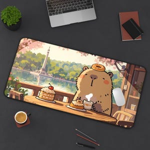 Op de afbeelding: Een rechthoekige muismat met een cartoonbeer die geniet van een maaltijd in een café met een schilderachtig uitzicht. De beer heeft een donut op zijn hoofd en zit aan een tafel met cake en koffie. De achtergrond toont een meer en bomen.