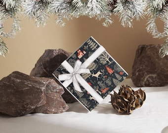 May include: A square gift box wrapped in dark navy paper featuring a repeating pattern of white deer, red houses, and evergreen trees. The box is tied with a white satin ribbon bow. The design includes small red circular accents and white foliage.