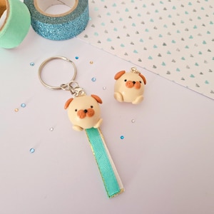 Peut inclure: Un porte-clés pug de couleur crème avec des oreilles et un nez bruns, attaché à un ruban turquoise et blanc. Un charm pug assorti est également présent. Des rouleaux de ruban adhésif washi décoratifs turquoise et pailletés sont en arrière-plan.