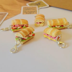 Puede incluir: Llaveros en miniatura con forma de sándwiches con rellenos de jamón, queso, lechuga y huevos. Cada llavero tiene un cierre plateado. Los sándwiches están sobre una superficie blanca.
