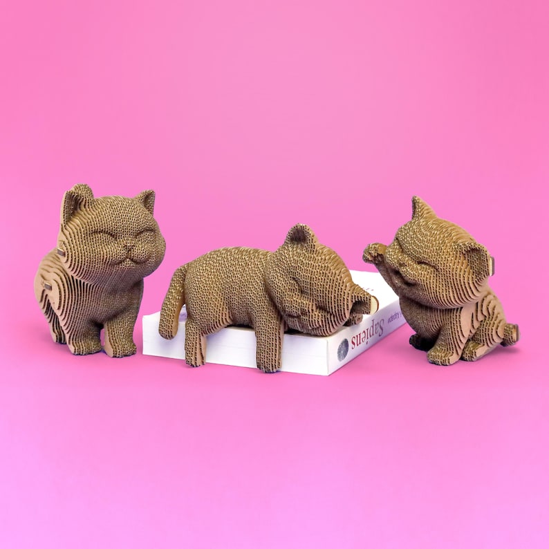 May include: Three cat figurines constructed from layered brown corrugated cardboard. One cat stands on the left, one lies across a white book titled Sapiens in the centre, and one sits on the right with a raised paw. All have closed eyes.