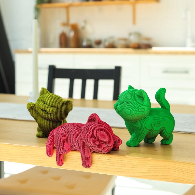 May include: Three small cat figurines with a layered, 3D-printed texture. One is olive green and sitting, one is bright green and standing, and one is magenta and lying down with its front paws hanging over the edge of a wooden surface.