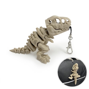 Puede incluir: Un dije de esqueleto de Tyrannosaurus Rex beige con un cordón negro y un cierre plateado. El dije de dinosaurio está sujeto a una bolsa negra en la esquina inferior derecha. El dije es una réplica en miniatura de un esqueleto de T-Rex.