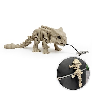 Puede incluir: Figura de esqueleto de dinosaurio beige con un cordón negro y un llavero plateado. La figura es una réplica detallada de un Ankylosaurus. Una versión más pequeña de la figura está sujeta a una mochila negra en un recuadro circular.