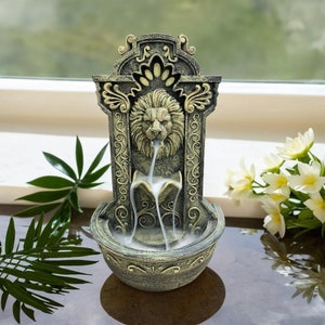 Op de afbeelding: Een decoratieve wierookbrander in de vorm van een fontein met een leeuwenkop. De wierookrook stroomt uit de leeuwenbek naar beneden en creëert een waterval-effect. De fontein is gemaakt van grijs en gouden keramiek.