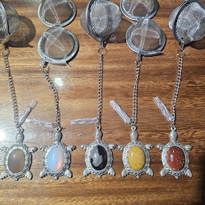 Könnte beinhalten: Sechs silberne Tee-Infuser mit Ketten und Schildkröten-Anhängern. Jede Schildkröte hat einen andersfarbigen Stein in ihrem Panzer: braun, weiß, schwarz, gelb und rot.