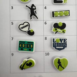 May include: Ten tennis shoe charms featuring various tennis-themed designs, including a tennis player, a tennis racket, a tennis ball, a heart with the word "Tennis" on it, a scoreboard, a tennis bag, and a heart with a tennis player inside. The charms are all white with green and black accents.