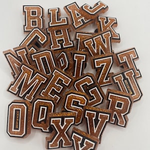 May include: A collection of brown glitter letters with white outlines. The letters are in a variety of sizes and are arranged in a random order. The letters are all uppercase and include the letters A, B, C, D, E, F, G, H, I, J, K, L, M, N, O, P, Q, R, S, T, U, V, W, X, Y, and Z.