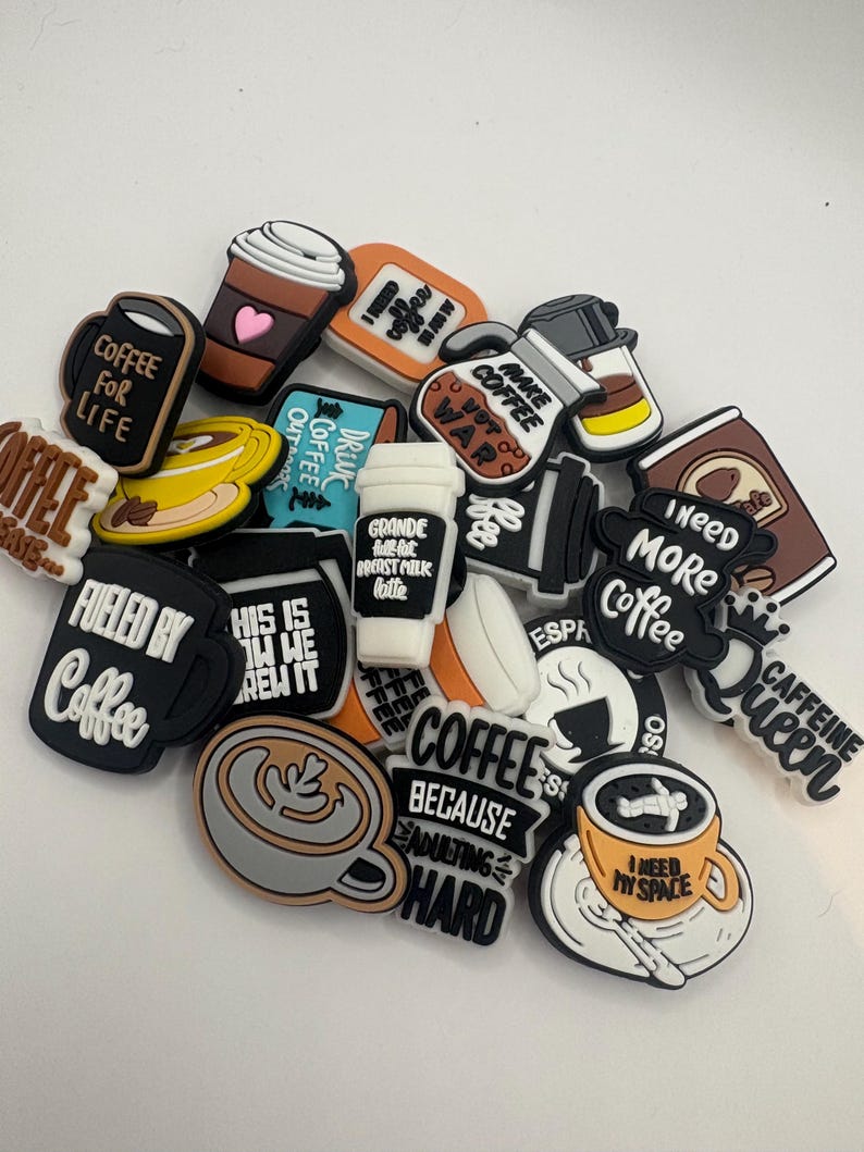 May include: A collection of coffee-themed charms in various shapes and colors. The charms feature coffee cups, coffee pots, and phrases like "Coffee for Life," "Fueled by Coffee," and "I Need More Coffee." The charms are designed for decorative purposes.