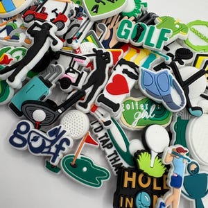Könnte beinhalten: Eine Sammlung bunter Golfschuh-Anhänger. Die Anhänger zeigen verschiedene golfbezogene Designs, darunter Golfbälle, Golfschläger, Golfwagen und die Worte "Golf" und "Hole in One".
