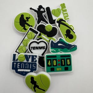 May include: A collection of green, black and white tennis shoe charms. The charms feature tennis-themed designs, including tennis rackets, tennis balls, and the words "Love Tennis Championship".