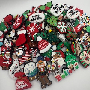 May include: A pile of festive Christmas charms in various shapes and sizes. Designs include Santa Claus, Christmas trees, and snowmen, with text like "Happy New Year" and "Merry Christmas." The charms are predominantly red, green, and white, with other seasonal colours.