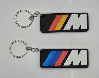 May include: Two black keyrings with a silver ring. The keyrings feature the BMW M logo in white with coloured stripes. One keyring has red, yellow, and blue stripes, while the other has blue, red, and white stripes.