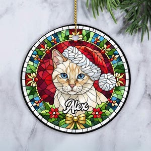 May include: A round Christmas ornament featuring a cat wearing a Santa hat, designed in a stained-glass style. The cat has blue eyes and the name "Alex" is written below. The ornament has a gold string for hanging and is surrounded by a colorful border.