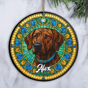 May include: A round ceramic ornament featuring a brown dog with a light blue collar, centered against a vibrant stained glass pattern of yellow and blue leaves. The name Alex is printed in white script at the bottom, with a gold hanging cord.