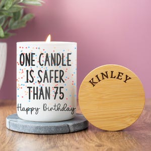 May include: A white candle with the text "ONE CANDLE IS SAFER THAN 75 Happy Birthday" and colorful confetti dots. A wooden lid with "KINLEY" is next to the candle. The candle is on a gray stone coaster.
