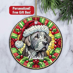 May include: A stained glass Christmas ornament featuring a black Labrador Retriever wearing a red and white Santa hat. The ornament has a green holly wreath around the dog and the name "Franko" written in a festive font.