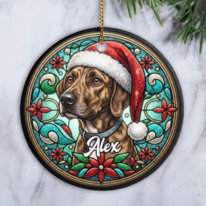 May include: Round Christmas ornament featuring a brown dog wearing a Santa hat. The dog's name, "Alex", is printed on the collar. The ornament has a stained-glass style design with red and green floral accents and a gold border. A gold string is attached for hanging.