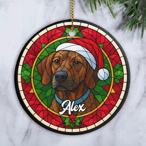 May include: Round Christmas ornament featuring a brown dog wearing a Santa hat and a blue collar. The dog's name, Alex, is written in cursive. The ornament has a stained-glass design with red, green, and gold accents, and a black border.