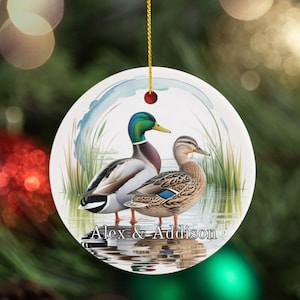 May include: Round Christmas ornament featuring a watercolor painting of two ducks in a pond. The male duck has a green head and the female is brown. The ornament has the names "Alex & Addison" printed on it. A gold string is attached for hanging.