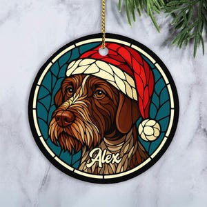 May include: Round Christmas ornament featuring a brown and white dog wearing a red and white Santa hat. The dog's name, Alex, is written in cursive. The ornament has a gold hanging string and a teal background.