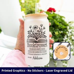 May include: A frosted glass jar with a wooden lid and straw, featuring the word "Booktrovert" and a floral design. The jar includes the name "Addison" and "Charlotte" on the lid. The jar is held by a person.
