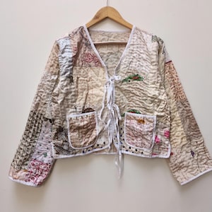 May include: A patchwork jacket with a V-neck and tie closure. The jacket features a variety of fabric patterns in beige, brown, and red hues. It has two front pockets and white trim. The jacket is hanging on a wooden hanger.