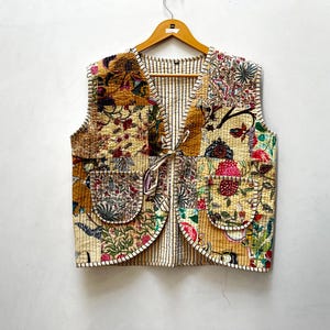 Peut inclure: Un gilet matelassé avec un motif patchwork présentant des motifs floraux et botaniques dans des tons or, rouge et vert. Le gilet a deux poches avant et une doublure intérieure rayée. Il est suspendu à un cintre en bois.