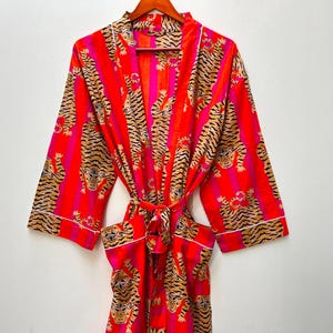 May include: A vibrant red robe with a tiger print and pink stripes. The robe features a tie waist, white trim, and a repeating pattern of tigers in shades of brown and black. The robe is hanging on a wooden hanger.