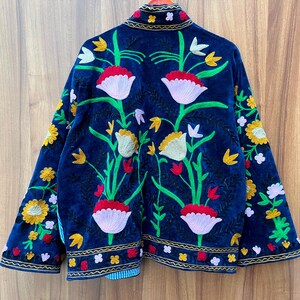May include: A dark blue jacket with vibrant floral embroidery. The jacket features a detailed design of red, white, yellow, and green flowers and leaves. The edges are trimmed with gold and the jacket is made of a soft, velvety material.