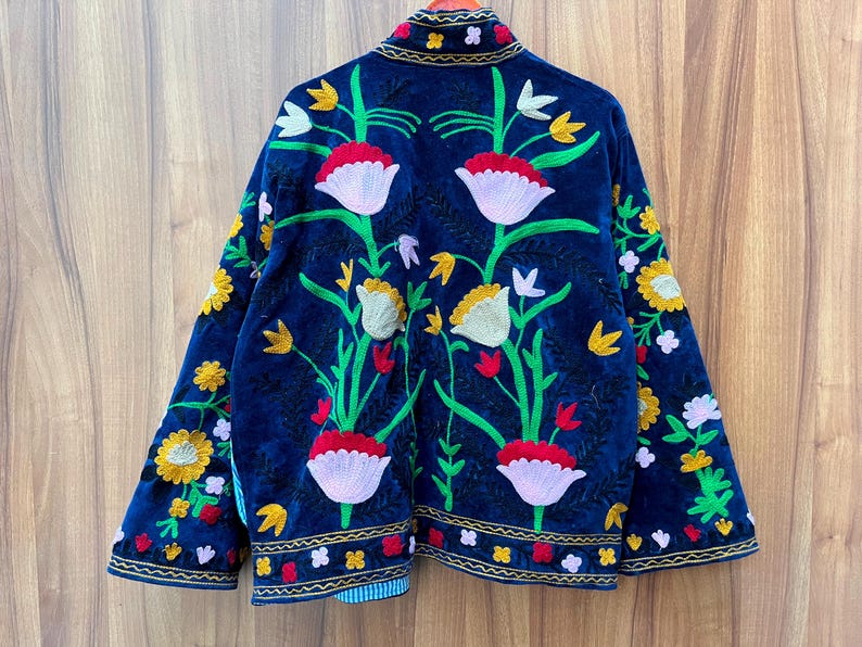 May include: A dark blue jacket with vibrant floral embroidery. The jacket features a detailed design of red, white, yellow, and green flowers and leaves. The edges are trimmed with gold and the jacket is made of a soft, velvety material.