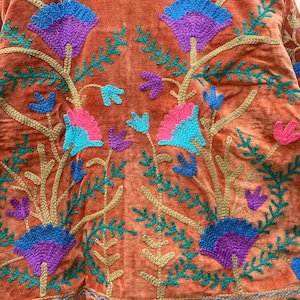 May include: Close-up of a rust-colored velvet jacket with intricate floral embroidery. The embroidery features blue, purple, and pink flowers with green leaves and gold stems. The jacket's design is symmetrical and detailed.