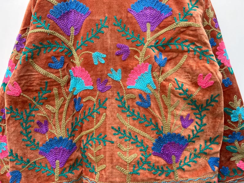 May include: Close-up of a rust-colored velvet jacket with intricate floral embroidery. The embroidery features blue, purple, and pink flowers with green leaves and gold stems. The jacket's design is symmetrical and detailed.