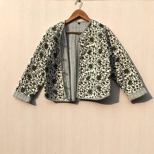 Op de afbeelding: Een wit gewatteerd jack met een groene bloemenprint. Het jack heeft een knoopsluiting en lange mouwen.