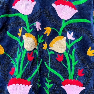 May include: A close-up of a navy blue jacket with intricate floral embroidery. The design features large pink and white flowers, yellow accents, and green stems. The embroidery is detailed and adds texture to the garment.