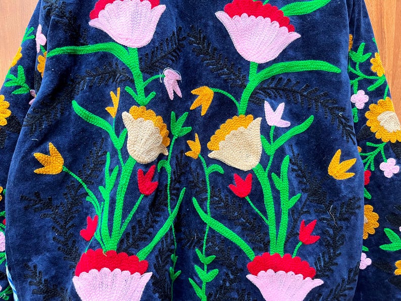 May include: A close-up of a navy blue jacket with intricate floral embroidery. The design features large pink and white flowers, yellow accents, and green stems. The embroidery is detailed and adds texture to the garment.