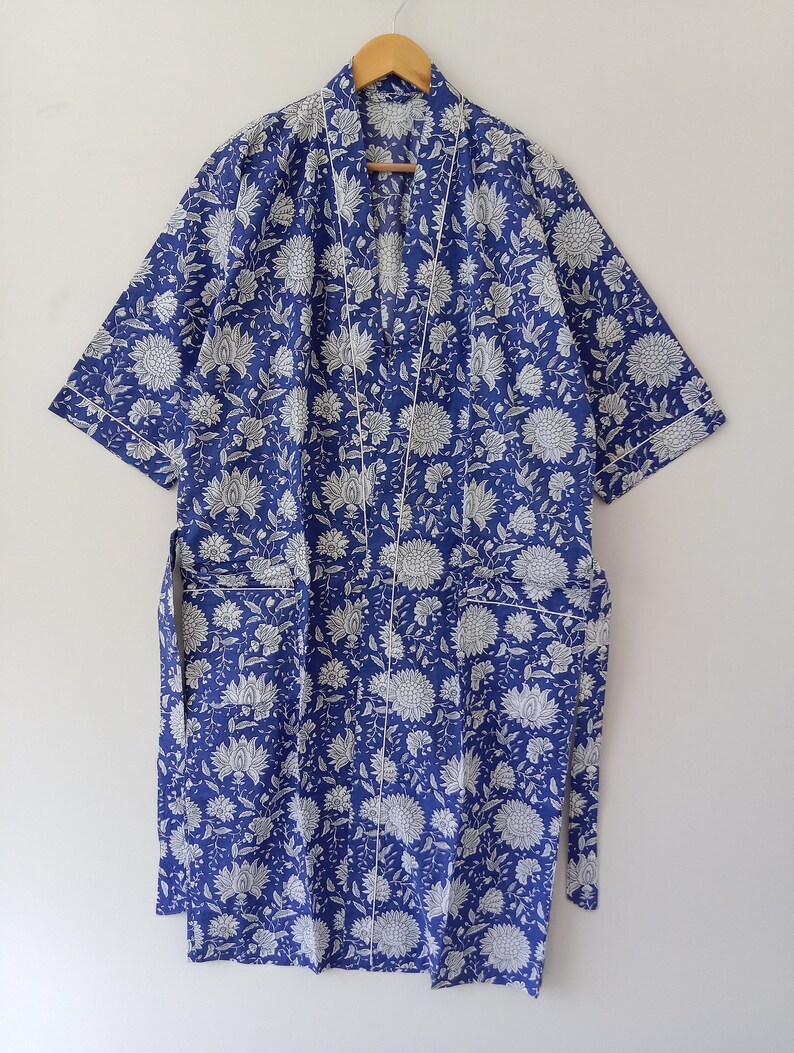 Op de afbeelding: Een blauwe katoenen badjas met een witte bloemenprint en witte bies. De badjas heeft een strikceintuur en twee zakken.