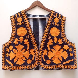Op de afbeelding: Een marineblauwe vest met oranje bloemenborduurwerk. De vest heeft een gestreepte voering en wordt gepresenteerd op een houten hanger. Het borduurwerk toont grote bloemen en gedetailleerde bladerpatronen, waardoor een decoratief kledingstuk ontstaat.