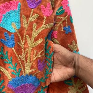 May include: Close-up of a rust-colored jacket with intricate floral embroidery. The embroidery features vibrant colors including pink, blue, purple, and green. The jacket has a pocket, and the design is detailed with gold-colored thread.