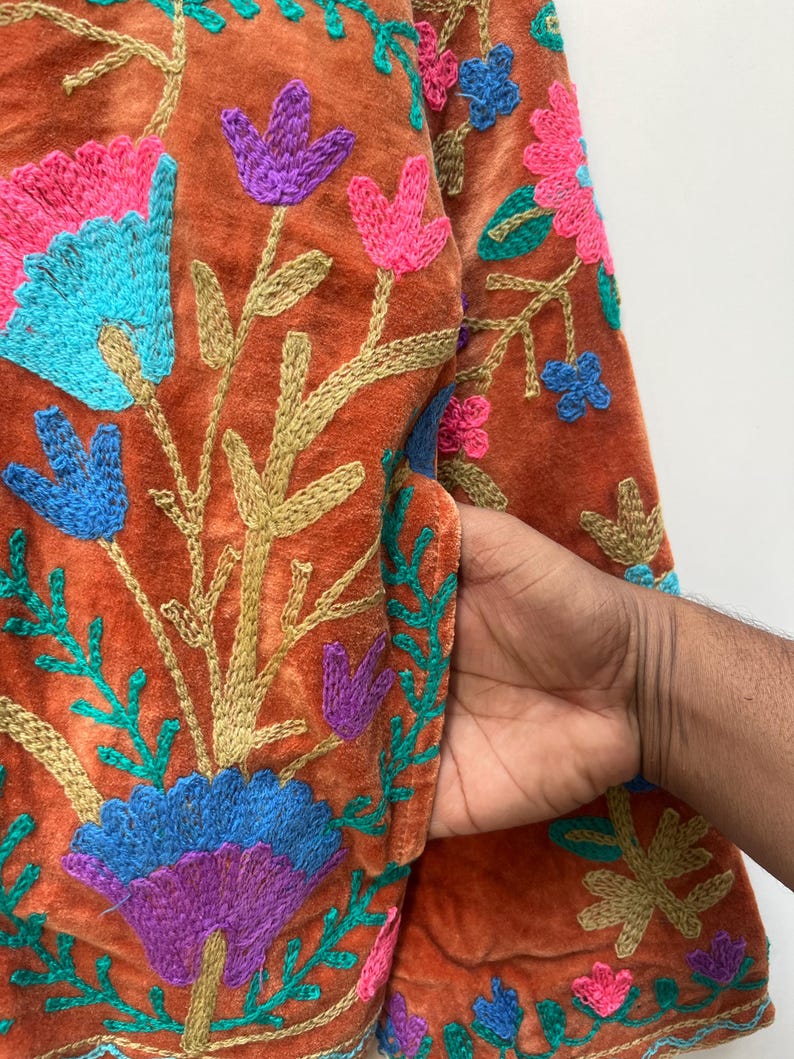 May include: Close-up of a rust-colored jacket with intricate floral embroidery. The embroidery features vibrant colors including pink, blue, purple, and green. The jacket has a pocket, and the design is detailed with gold-colored thread.
