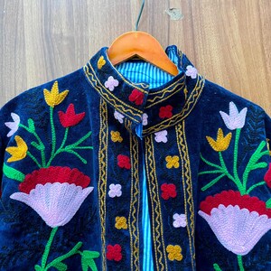 May include: A dark blue velvet jacket with colorful floral embroidery. The jacket features red, yellow, and white flower designs with green stems and leaves. The collar and edges are trimmed with gold and the lining is striped.