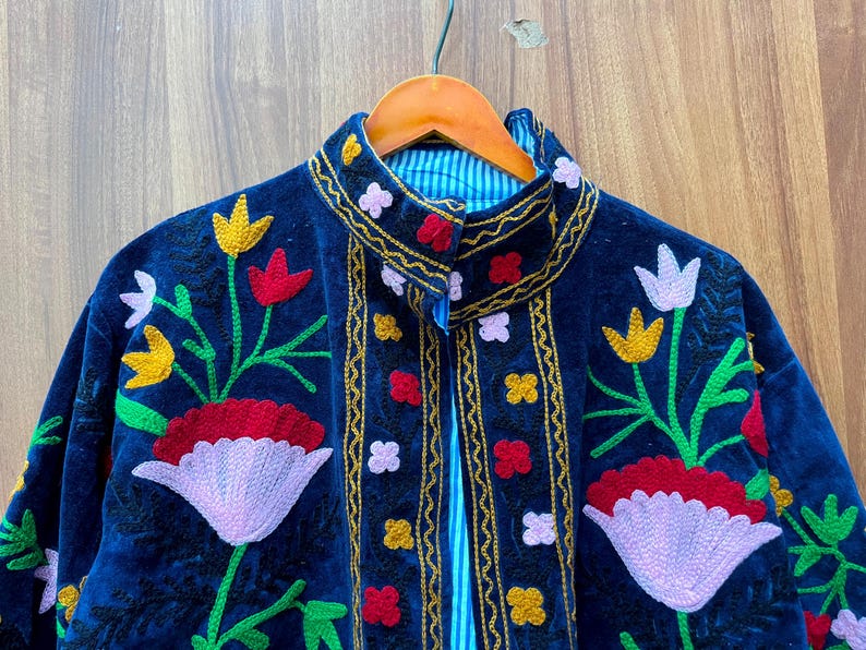May include: A dark blue velvet jacket with colorful floral embroidery. The jacket features red, yellow, and white flower designs with green stems and leaves. The collar and edges are trimmed with gold and the lining is striped.