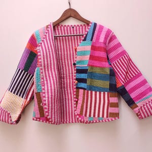 May include: A vibrant patchwork jacket with a V-neck and long sleeves. The jacket showcases a mix of fabrics in pink, purple, blue, green, and yellow. The interior lining is a pink and white striped fabric. The jacket is suspended from a wooden hanger.